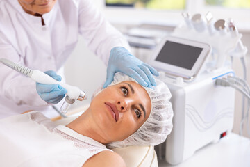 Concentrated young female patient undergoing facial procedure with application of atomized nutrients to enhance skin vitality at aesthetic medicine clinic. Modern technologies in hardware cosmetology