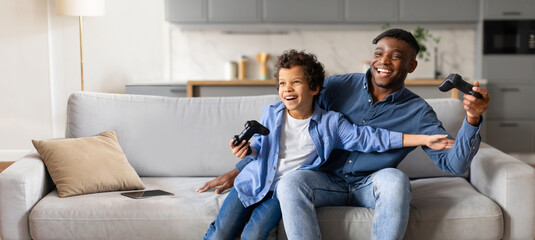 African American father and son share laughter while playing video games together on a comfortable sofa. The bright living room is filled with warmth and joy during their bonding time.