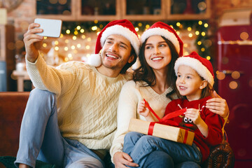 Young cheerful family in Santa hats with gifts taking selfie on smartphone in cozy kitchen on Christmas eve, panorama