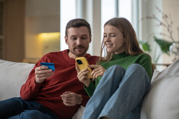 Happy couple online shopping at home sits on comfortable couch using phone and bank card. Interested satisfied woman holding smartphone making internet purchases with man husband. Spending money.
