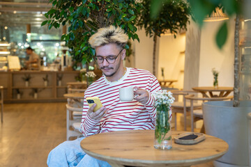 Thoughtful young man drinking coffee at cafe, using smartphone. Guy take break from work, chatting...