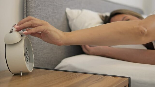 Woman turns off alarm clock in early morning, waking up slowly. Morning light softly fills room as she stretches and starts her day. Perfect clip to depict morning routines or daily wake-up habits.