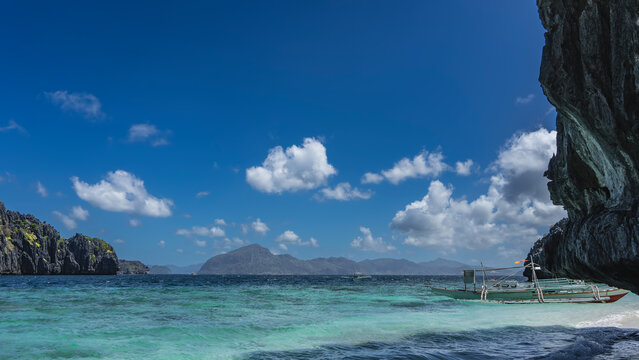 The traditional Filipino bangka boat is moored off the shore of the aquamarine ocean. A sheer cliff in the foreground. Mountains in the distance against the blue sky and clouds. Philippines. Palawan.