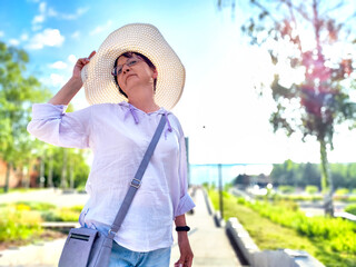 A woman is playfully adjusting her large sun hat while standing in a vibrant park filled with trees under a clear blue sky