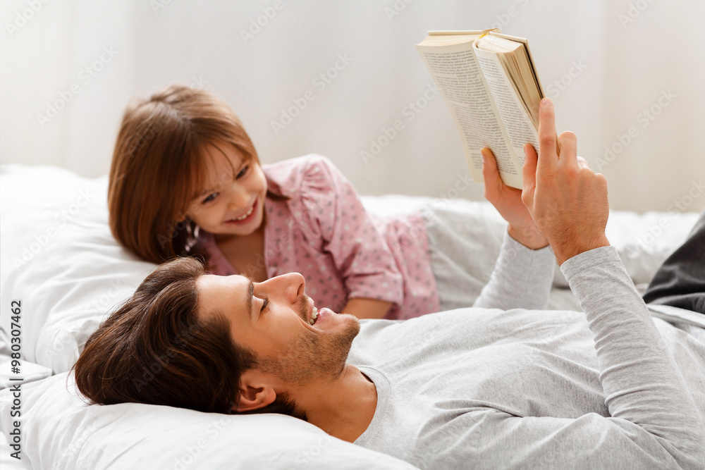 Wall mural Young father reading book to his little cheerful daughter, laying on bed together