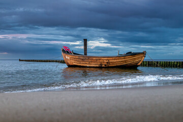 Fototapeta premium Tranquil Coastal Scene Featuring a Lonely Anchored Fishing Boat Against Blue Clouds at the Blue Hour, with Waves Gently Hitting the Beach and Groynes Along the Shore