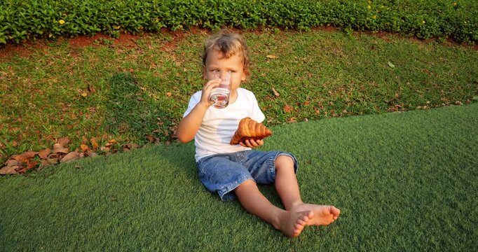 Young boy enjoys snack outdoors, sitting on artificial grass during mini picnic. He takes sip of water from glass, then bites into end of large croissant, glancing at camera.