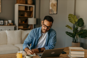 A busy, focused young adult male freelancer sitting at a desk and working on a project on his laptop.