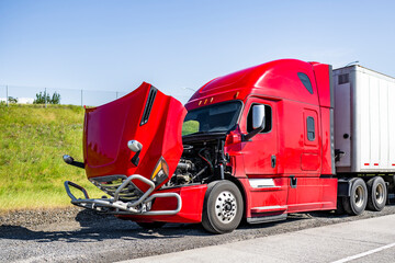 Broken red big rig semi truck with open hood and dry van semi trailer standing out of service on the road shoulder waiting for the towing truck or mobile road assistant for engine repair help