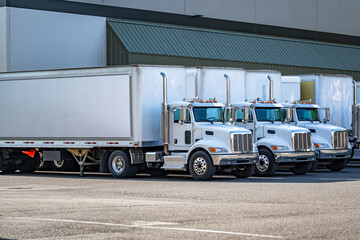 Day cab white big rig semi trucks with dry van semi trailers standing in row on warehouse parking...