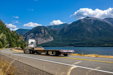 Long haul white big rig semi truck tractor with empty step deck semi trailer going to warehouse for...