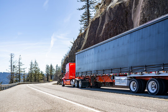 Low cab profile red big rig semi truck transporting cargo in flat bed conestoga semi trailer driving on the mountain road with rock cliff and abyss on the sides