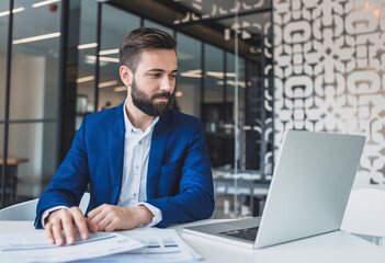 Happy young man working in the office with documents, seated at a desk with a laptop, conducting an online meeting with clients and partners while signing agreements and invoices.