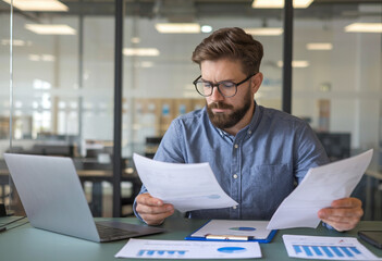 Fototapeta premium Stunned young office worker seated at a table, looking seriously surprised at the documents and bills he's holding.