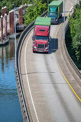 Team of two big rig semi trucks transporting cargo in green containers running on the turning overpass road along the Willamette River in Portland