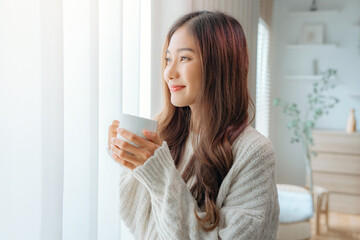 Attractive lady enjoying her morning coffee or tea at home, Smiling woman holding cup of tea in hands while looking out the window at morning.