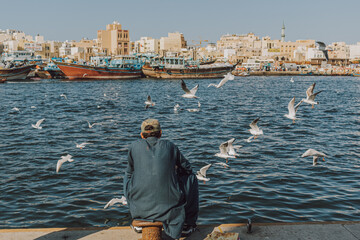 Dubai, UAE - January 6, 2020: Man enjoying waterfront view with boats and feeding seagulls at Dubai Creek with panoramic cityscape