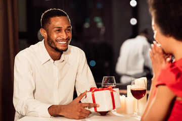 Obraz premium Cheerful Man Giving Wrapped Gift Box To Girlfriend Congratulating Her On Anniversary Having Romantic Date In Restaurant. Selective Focus