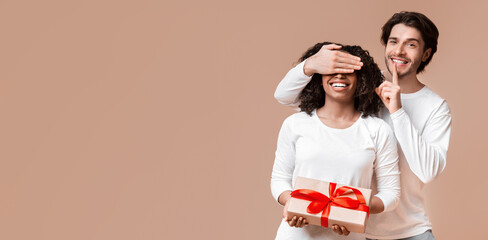 Guess who. Handsome guy covering his girlfriend's eyes, giving her present and making shhh gesture on yellow background with free space