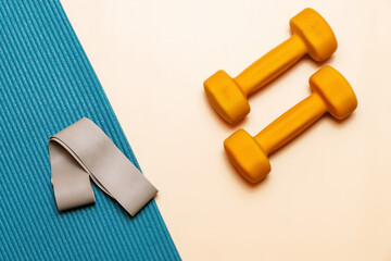 Pair of yellow dumbbells on a peach background. Yoga mat with a elastic band. The colors are...