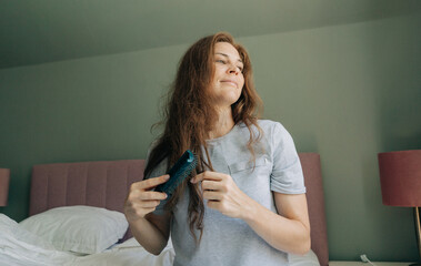 A woman sitting on a bed combs her long red hair.