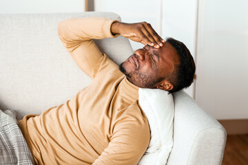 Afro Guy Having Severe Headache Touching Forehead Lying On Sofa At Home. Stressed Life Concept