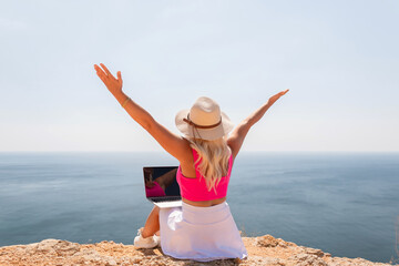 A woman is sitting on a rock overlooking the ocean with a laptop in front of her