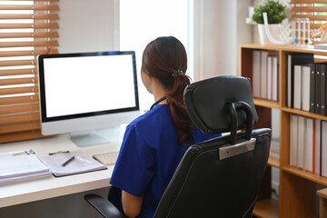 Back view of female doctor in blue scrubs sitting at desk in front of a computer monitor