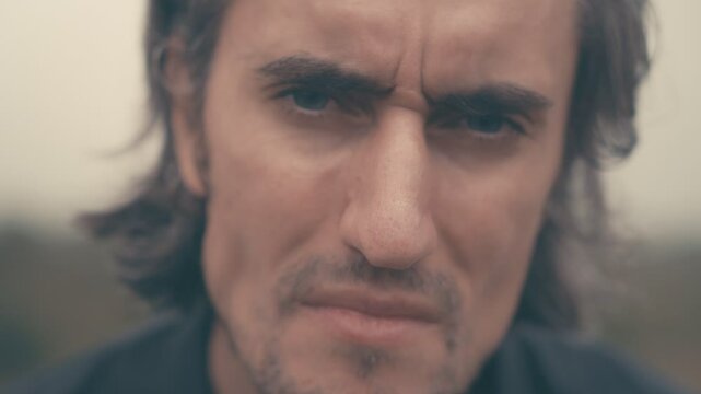 Young man of European appearance in a black shirt with long black hair, unshaven, adjusts hair, approaches, looks disdainfully at camera, nods, sighs, disapproves, smiles, close-up.