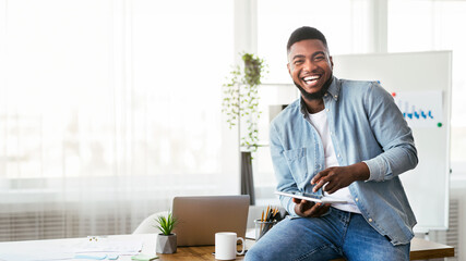 Work with fun. Portrait of cheerful black employee using digital tablet in modern office and laughing out loud