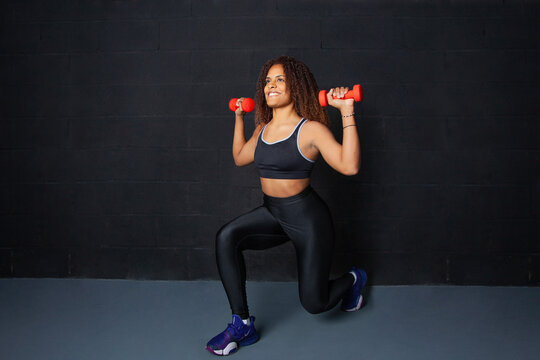 Chica afroamericana de 30 a&ntilde;os haciendo ejercicio de estocadas con pesas rojas en un entorno deportivo negro. estilo de vida saludable