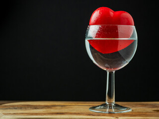 Red heart in a wine glass with water on wooden surface, dark background. Optical illusion, Concept of love and romance. Copy space.