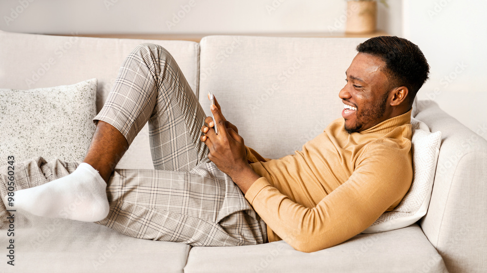 Poster mobile application. happy millennial afro guy using smartphone lying on couch at home.