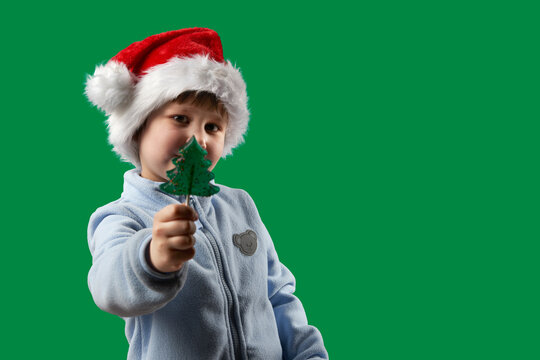 A child wearing a Santa Claus hat holds a lollipop on a stick in the shape of a Christmas tree in his outstretched hand. The background is green. Copy space.