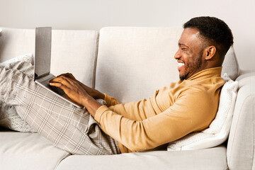 Happy African American Man Using Laptop Computer Lying On Couch Indoor. Weekend At Home Concept.