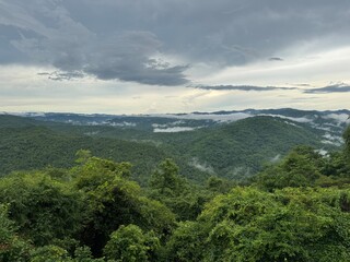 Obraz premium Expansive view of green mountains covered in mist, with dark clouds overhead creating a dramatic, atmospheric landscape. A perfect scene for themes of nature, wilderness, and adventure
