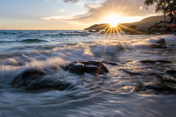 Naklejka premium Panoramic ultra wide landscape during sunset on the beach while waves crashing on the rocks in the coast and the sun's rays bright over the mountain in Koh Rong, Cambodia