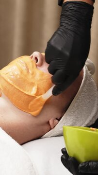 Close-up of a cosmetologist applying an alginate mask to a client's face with a spatula. The scene highlights professional facial skin care treatments in a beauty salon.