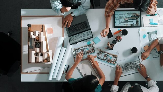 Top view of smart architect engineer writing at blueprint while sitting at meeting table with house model and laptop. Skilled manager working and taking about about building structure. Alimentation.