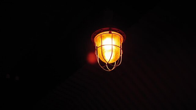 An explosion-proof lamp with a grille hangs from the ceiling in the tunnel of the bomb shelter. An empty scary dark corridor, a basement with concrete walls. Old electric lamps on the ceiling.