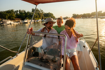 Family enjoying a boat ride on a sunset, father steering the boat, the mother sitting beside him, and their child standing between them.