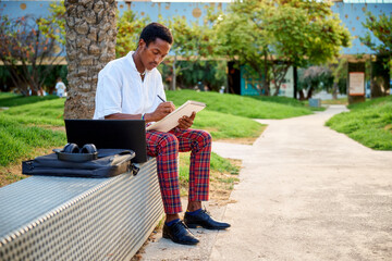 Fototapeta premium A young Black man sits on a bench in an urban area, sketching ideas in a notebook while his laptop and bag sit nearby