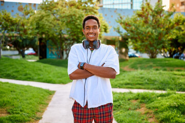 A young man smiles proudly with his arms crossed, wearing headphones, in a lively outdoor area