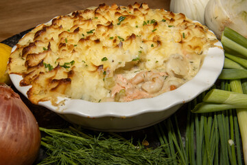 A delicious fish and seafood pie, topped with golden brown mash potato surrounded with the ingredients used to create the dish on a wooden kitchen table