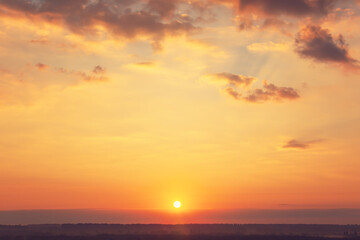 Sunset over the horizon. The sun sets beyond the trees, lighting the sky in warm shades of yellow, orange and red. The clouds, light and thin, create a pleasant contrast against the clear sky
