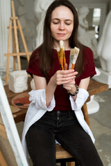 Female artist painter sits in studio holding few paintbrushes in hands