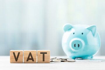 VAT is written on a wooden block beside a blue piggy bank and a pile of coins on a wooden table against a bright white background.  Financial planning, savings, and wise money management.