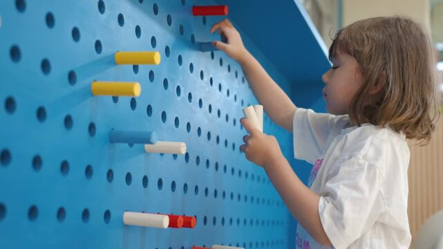 Little Girl Stick Wooden Multicolor Sticks into Peg Board Wall with Holes at Inside Playroom - a handheld close-up shot
