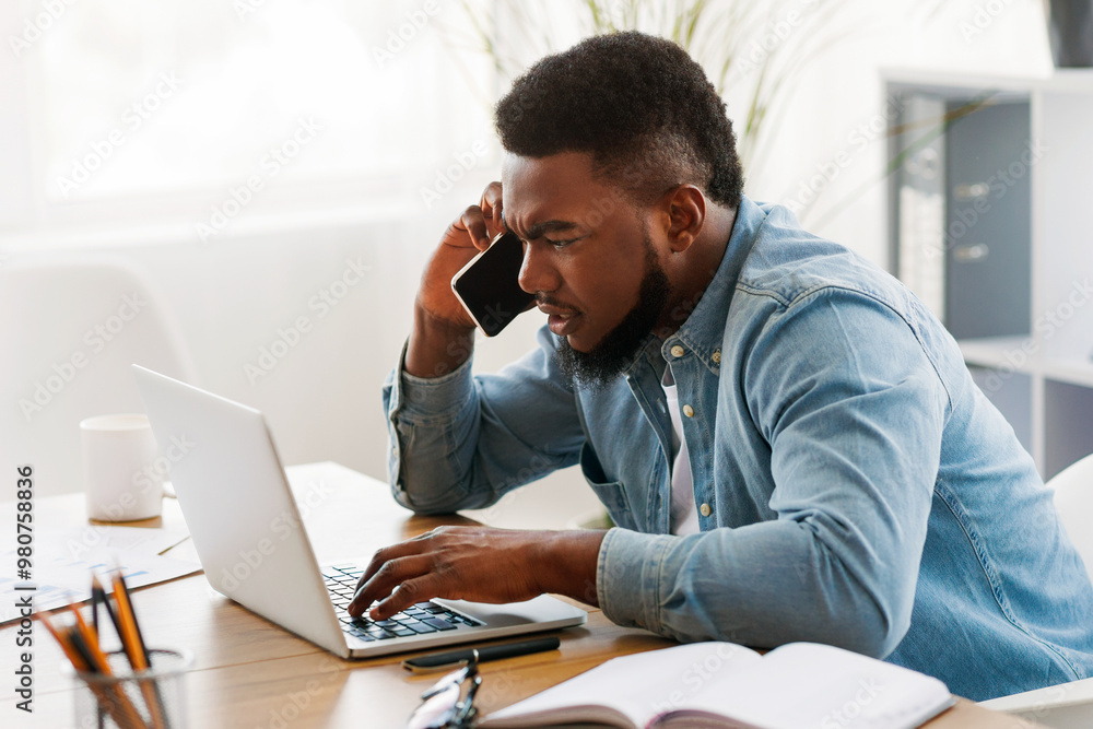 Poster worried black businessman talking on cellphone and looking at laptop screen, trying to solve problem