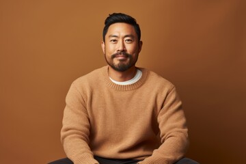 Obraz premium Portrait of a content asian man in his 30s wearing a cozy sweater while standing against soft brown background